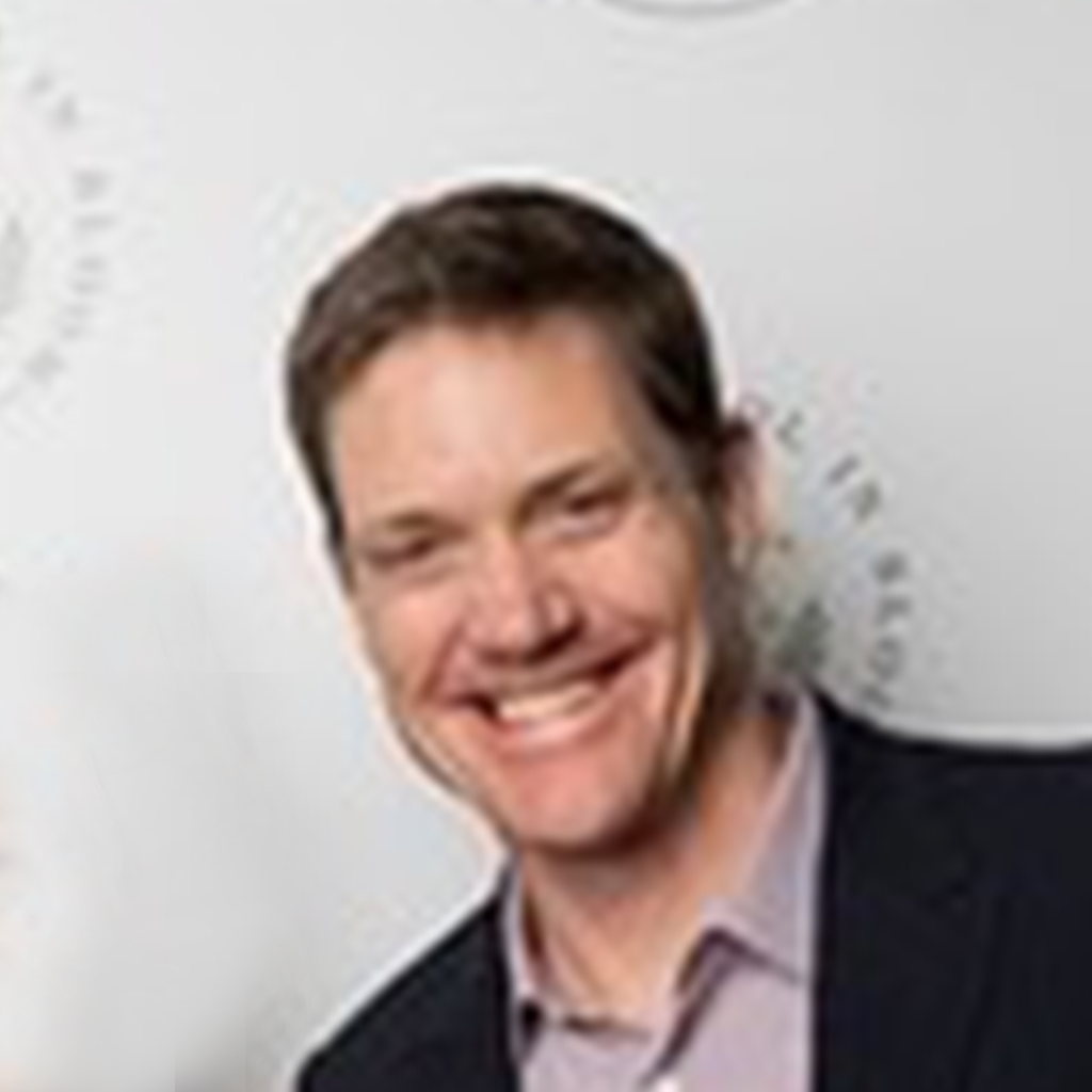 A man with short brown hair, wearing a dark jacket and light-colored shirt, smiles in front of a white background with circular text.