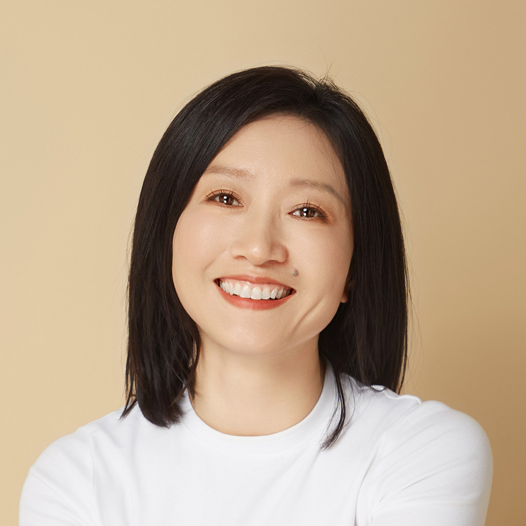 A woman with straight black hair and a white shirt smiles at the camera against a beige background.